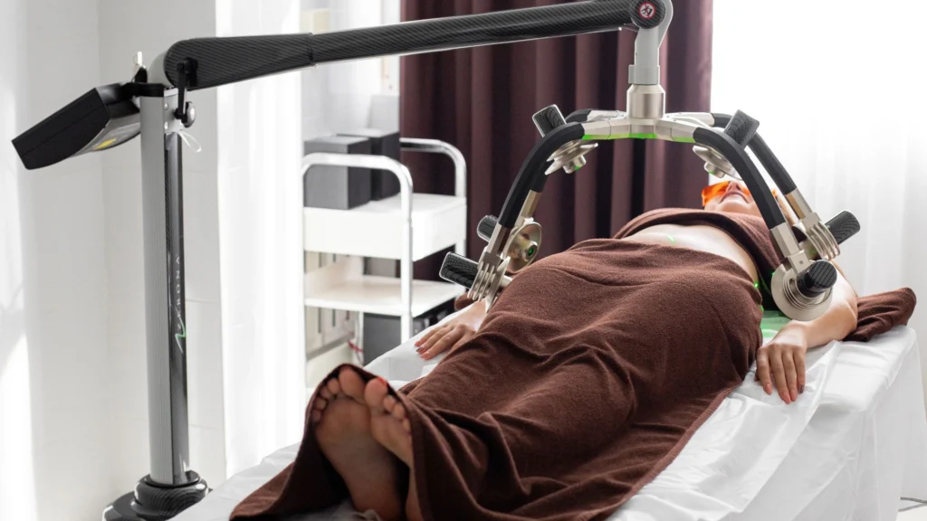 A patient receiving Class IV laser therapy using a robotic arm device in a modern clinical setting, wrapped in a brown towel, with soft lighting and medical equipment in the background.