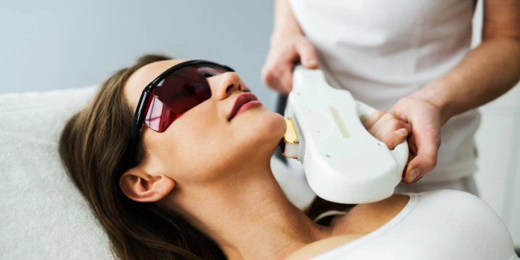 A woman receives facial laser therapy while wearing protective goggles during a non-invasive treatment session.