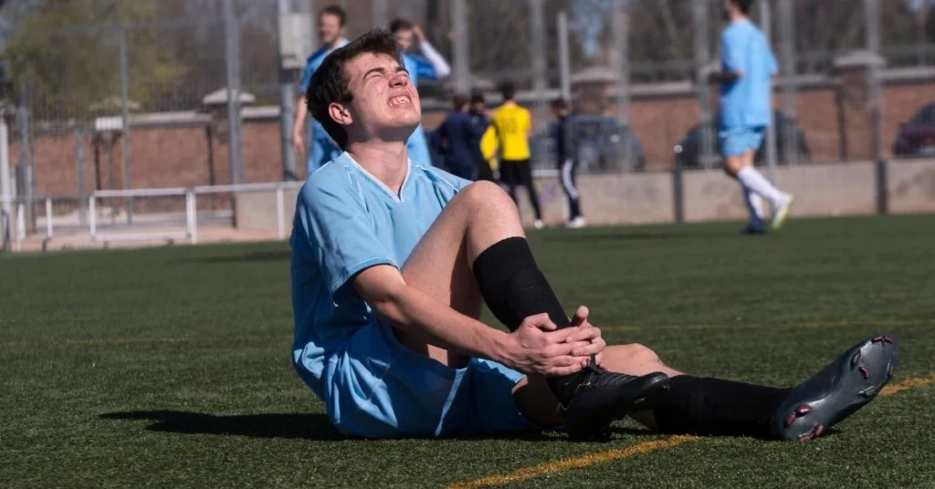 A-young-soccer-player-in-blue-uniform-sitting-on-the-field-holding-his-knee-in-pain-after-an-injury-during-a-match-with-other-players-in-the-background.